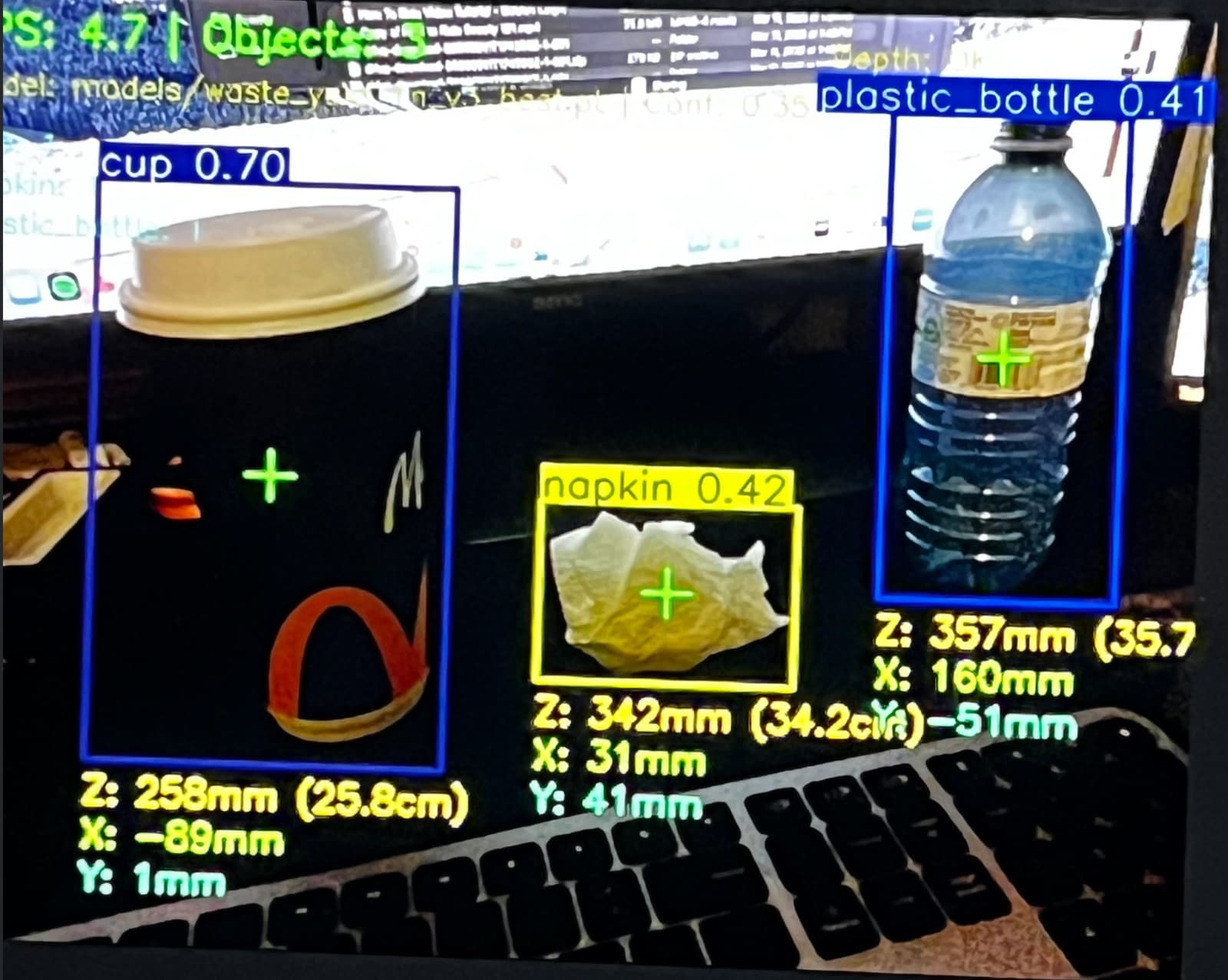 The RECLAIM perception node's live output: YOLO bounding boxes labeling a cup (0.70), a napkin (0.42), and a plastic bottle (0.41), each annotated with its 3D X/Y/Z position in millimetres from the depth camera.