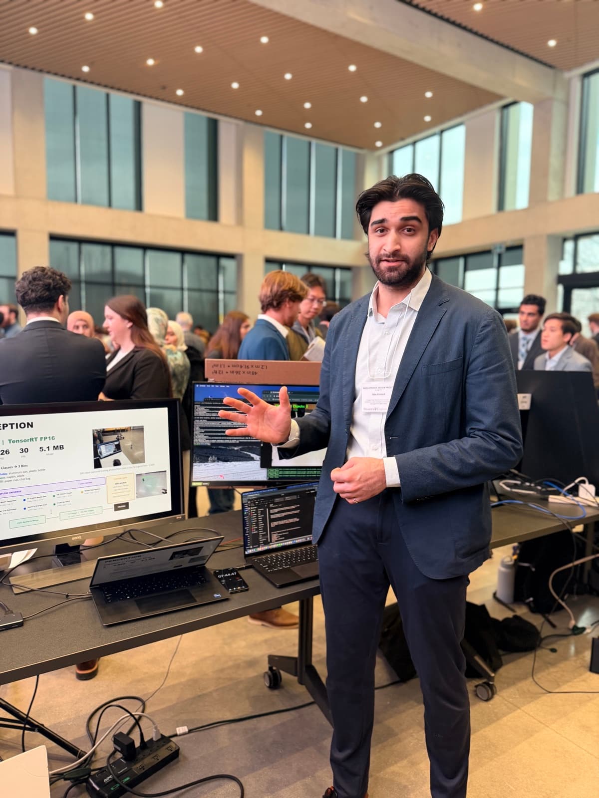 Issa presenting the RECLAIM perception stack at the showcase, monitors showing the detector's throughput, model size, and class-to-bin map.