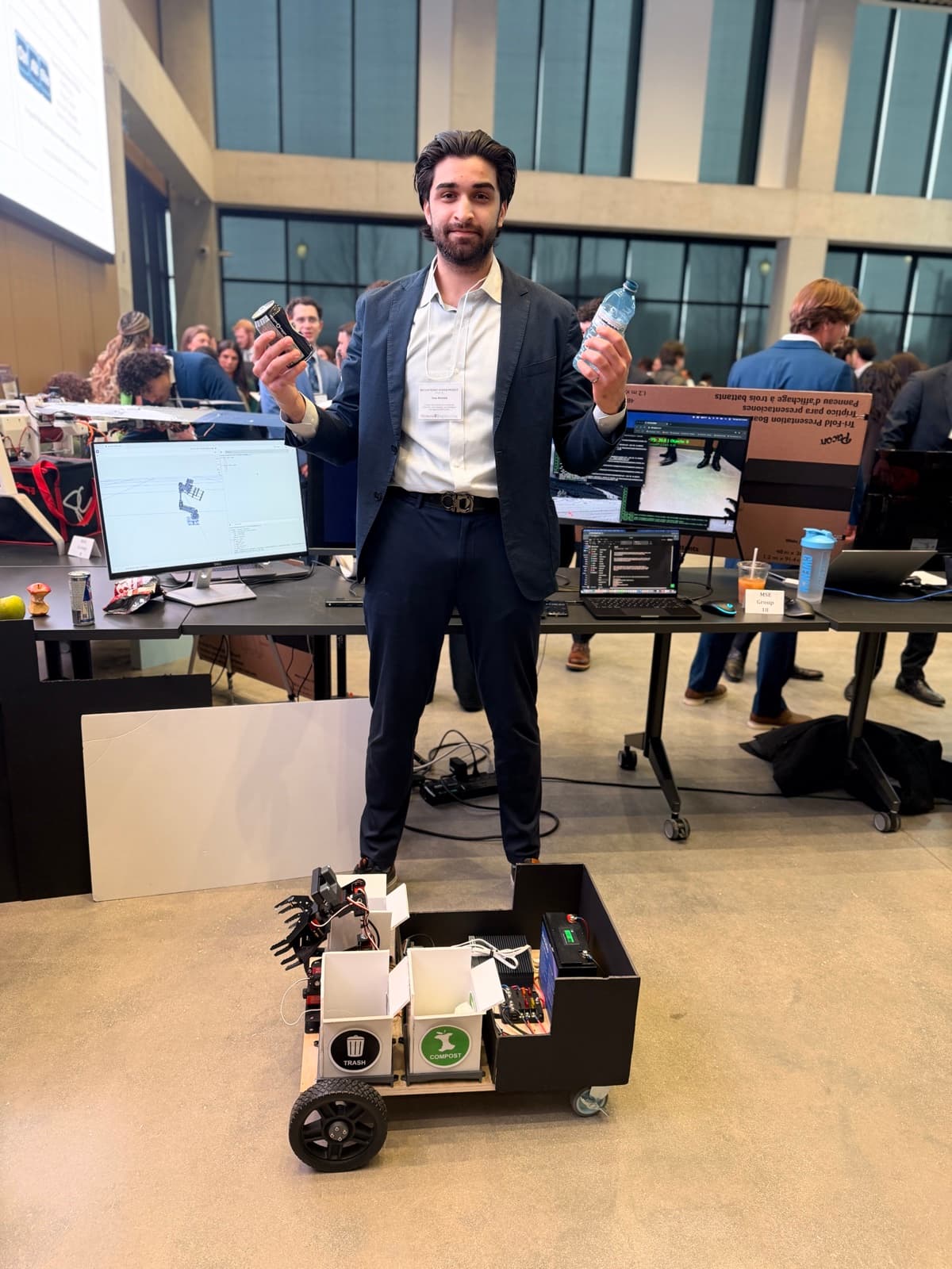 Issa Ahmed standing with the RECLAIM prototype at the showcase, holding an aluminum can and a plastic water bottle.